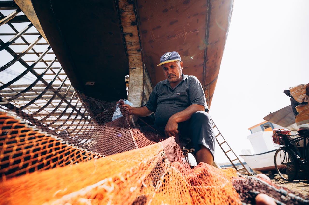 Fisherman repairs a net under a boat in Damietta, Egypt, highlighting traditional fishing techniques.