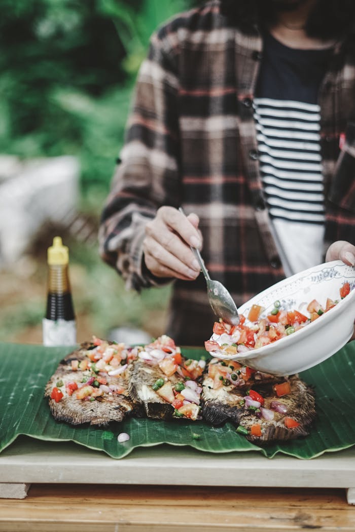 A person prepares a flavorful fish dish with fresh vegetables on banana leaves, adding vibrant toppings.